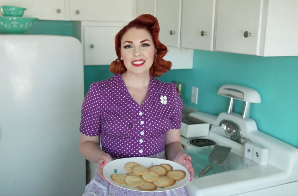 Luci fay wearing a 1950s purple colored dress and holding a white ceramic plate of cookies