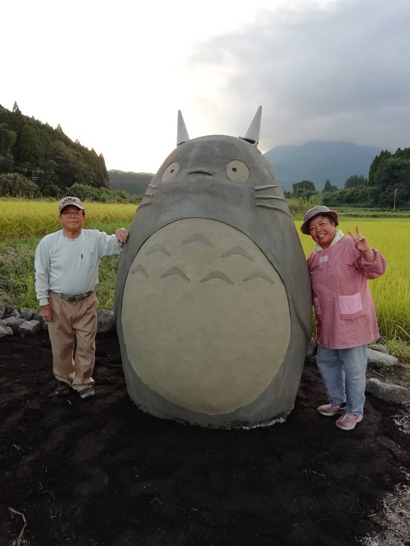 Two Japanese Grandparents Sculpted A Life-Size Totoro and standing next to it in the fields
