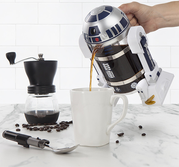 Black and grey-silver colored star wars themed coffee maker pouring black coffee in a white ceramic mug on a black-white marble surface; blacktop and transparent bottomed coffee  brewer with black coffee inside it with some coffee beans around it