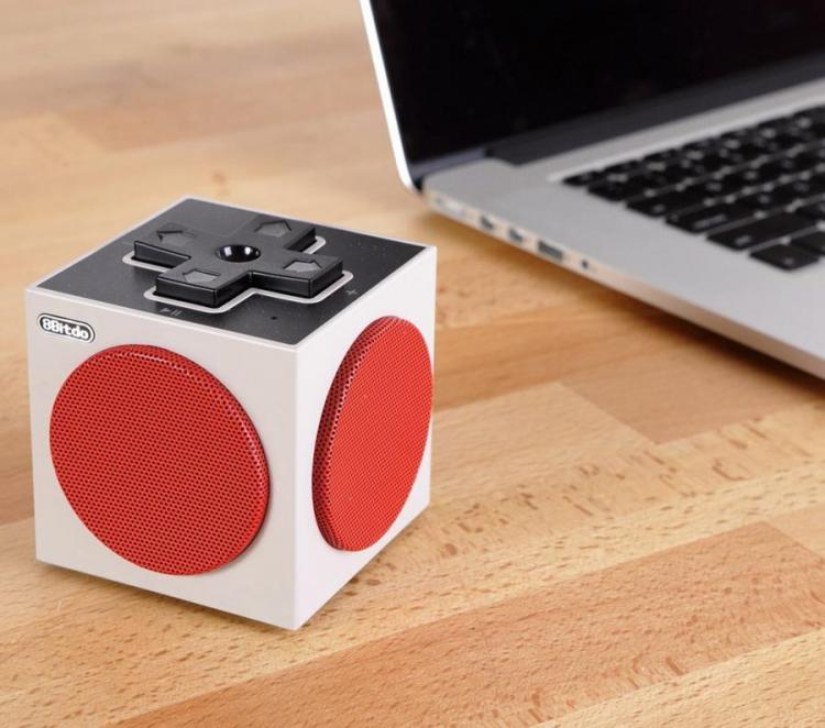 A white cubic Bluetooth speaker with red dots on every side and a black control top placed on a wooden table near a laptop