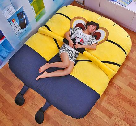A man sleeping on a giant blue and yellow minion themed bed on a wooden floor