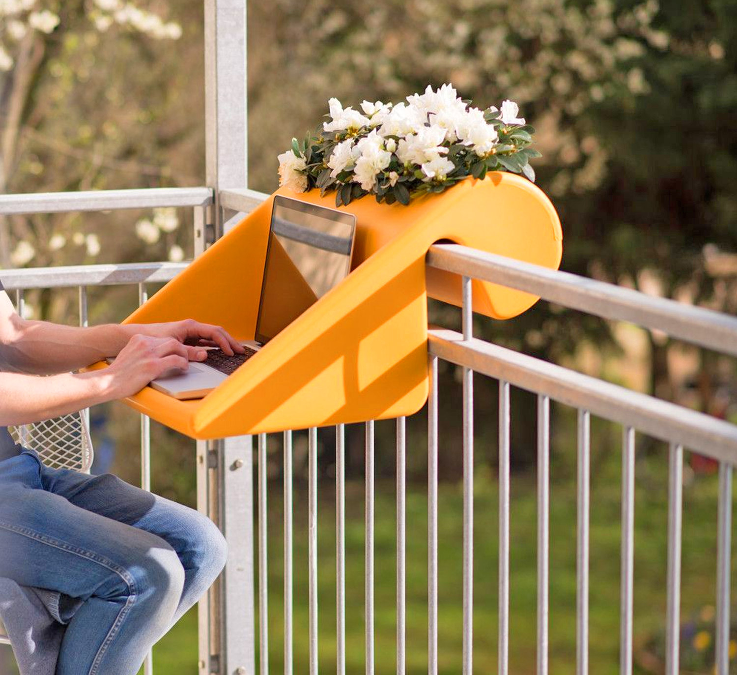 A girl working on a laptop on a yellow-colored mini balcony desk with a flower pot inside it