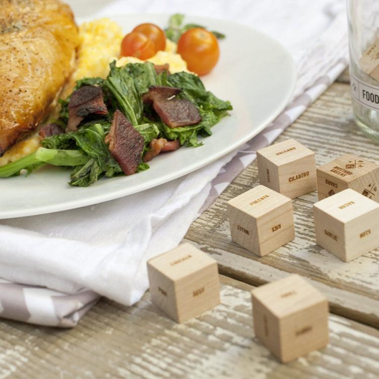 Wooden food decider dices on a wooden table near a food-filled white ceramic plate and a white cloth