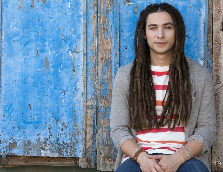 Jason Castro has a dreadlock hair style and behind him is a wood painted with sky blue color