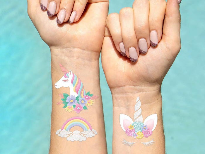 A girl having temporary unicorn-themed tattoos of a unicorn, a rainbow, and a unicorn horn on her arms against a blue background