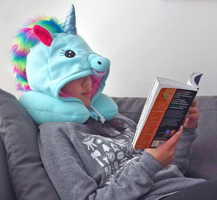 A girl, wearing a grey hoodie and hooded unicorn neck pillow, reading a book while resting on a grey sofa 