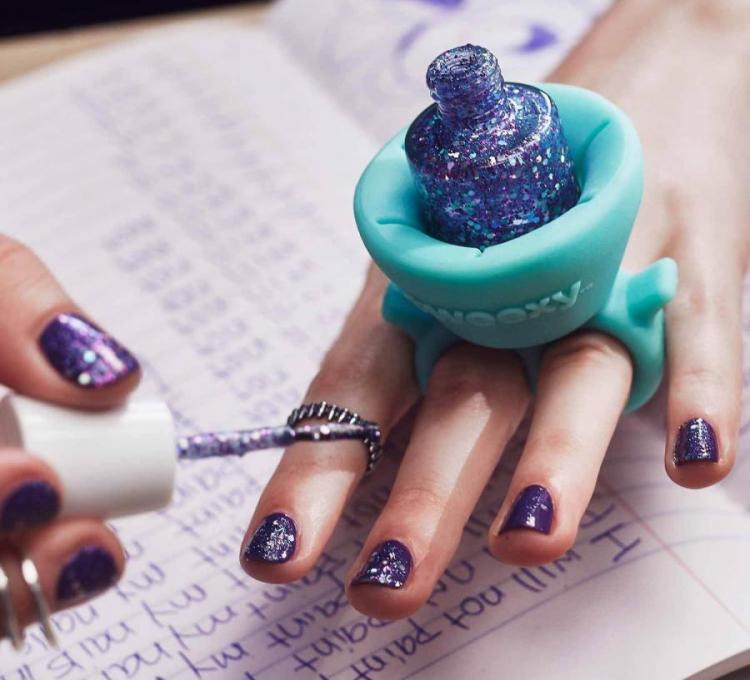 A girl applying shimmery blue-purple nail polish on her nails while holding a nail polish bottle in a light blue colored ring-like holder