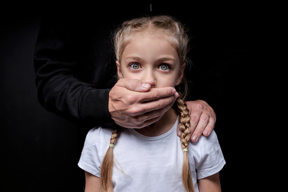In the dark, a man places his hands on the mouth of a young girl
