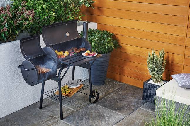 Black aldis smoker bbq, with food cooking inside it, in the grey marble floor backyard along with some plants