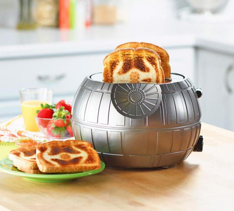 Grey colored star wars death star themed toaster with some toasted slices around it, a bowl of strawberries, and a glass of juice on a wooden kitchen tabletop