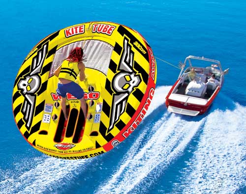 Blue shorts and a yellow life jacket-wearing man is on the giant kite tube which is yellow and black colored; a white boat with red details is pulling the kite tube in the ocean