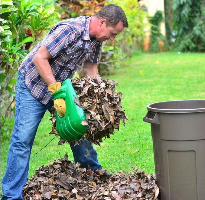 A man, wearing a blue and light blue checkered shirt, cleaning his garden with green colored giant claw leaf scooper