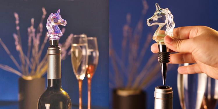 A LED glass unicorn-shaped wine bottle stopper on a bottle of wine in front of two wine glasses and a vase