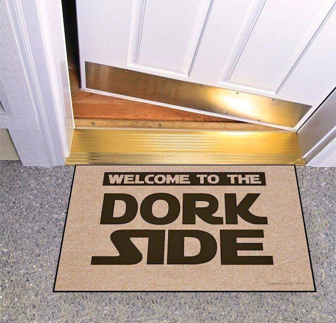 Black-brown 'Welcome To Dork Side' printed on a skin doormat on a grey marble floor next to a white-golden door