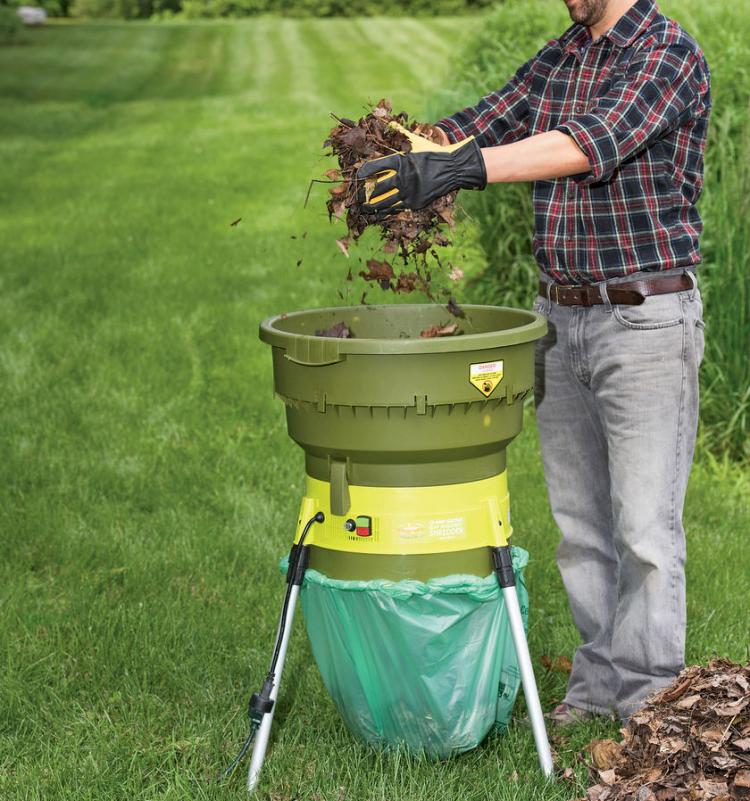 A man, wearing a black and pink check shirt and jeans, shredding leaves with the help of a green colored leaf shredder in a garden