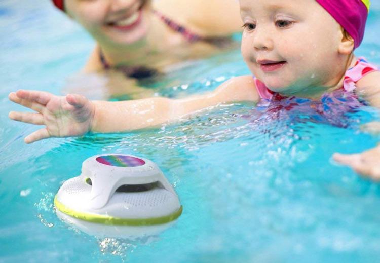 White and green colored Bluetooth speaker in the pool; a baby girl  and a girl enjoying the water