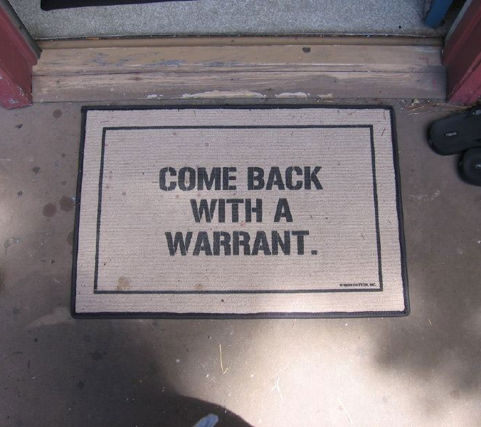 'come back with a warrant' printed on a white doormat with white details on a grey floor
