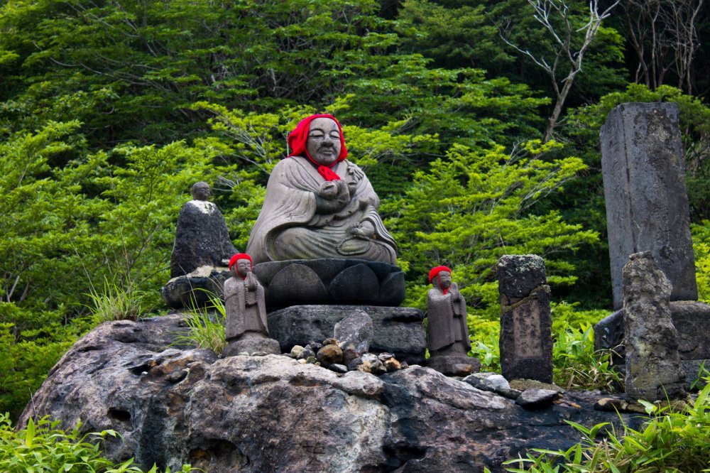 Black and grey colored giant and small statue wearing red colored hat on a stone in the forest
