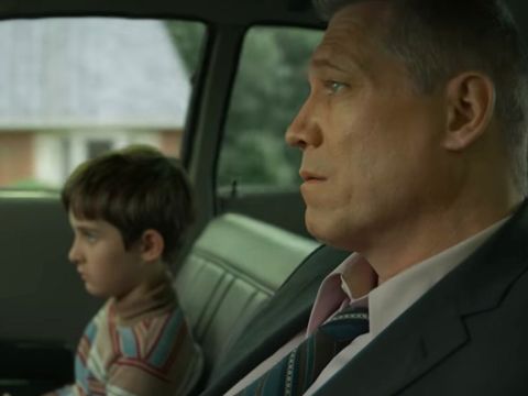 A man wearing black suit and a kid wearing a multi-colored sweater sitting in a car