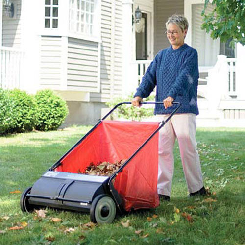 An old lady, wearing a blue sweater and white trousers, sweeping leaves with the red and black colored tool in garden