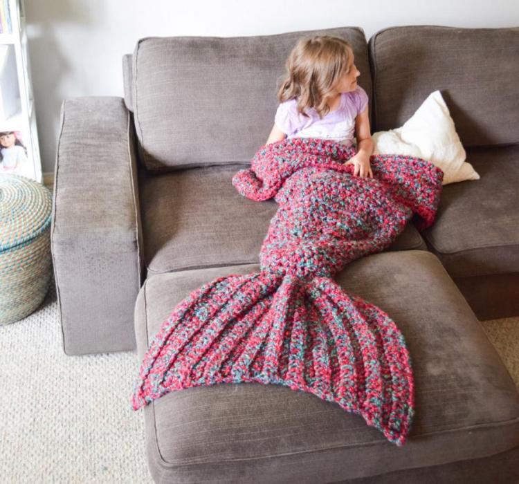 A girl wearing a pink and blue crocheted mermaid tail blanket while sitting on a grey sofa