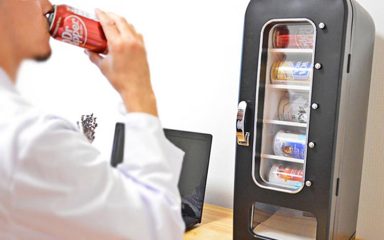 A man wearing a white shirt drinking can drink; a black Personal Mini Vending Machine with a black laptop on a wooden table