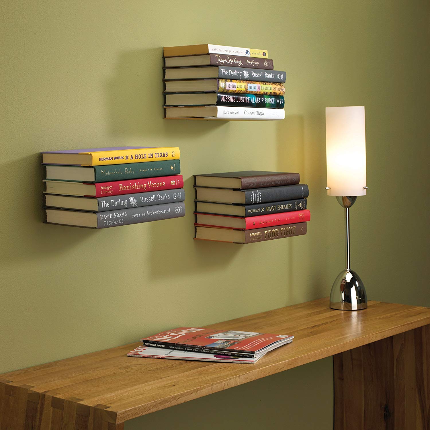 Three floating bookshelves on a henna colored wall beside a wooden table and a lamp
