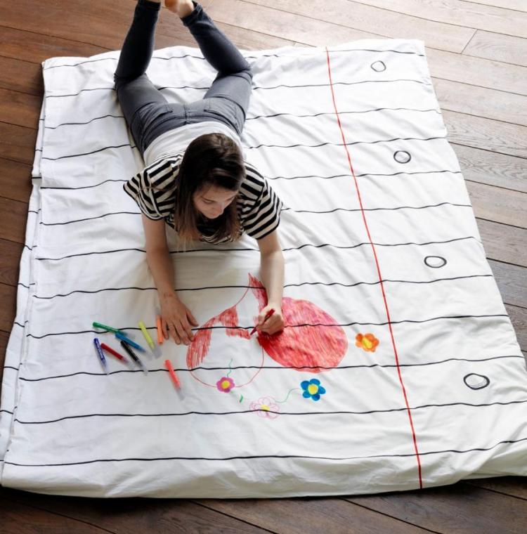 A girl lying and drawing on a white notebook page themed bed sheet on a brown wooden floor