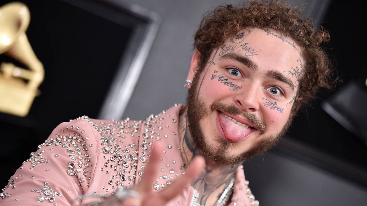 Post Malone with his weird face tattoos ,wearing a baby pink colored funky jacket, flaunting his tongue