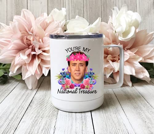White-colored nicolas cage printed mugs on a white-grey wooden top with some pastel pink flowers behind it