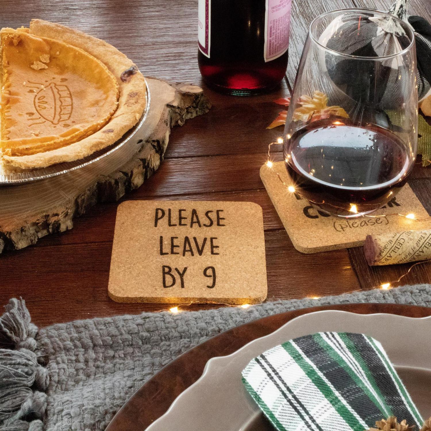 A square-shaped please leave by 9 coasters set on a wooden table along with half pumpkin pie in a pie pan and a less than a half-filled wine glass on the table