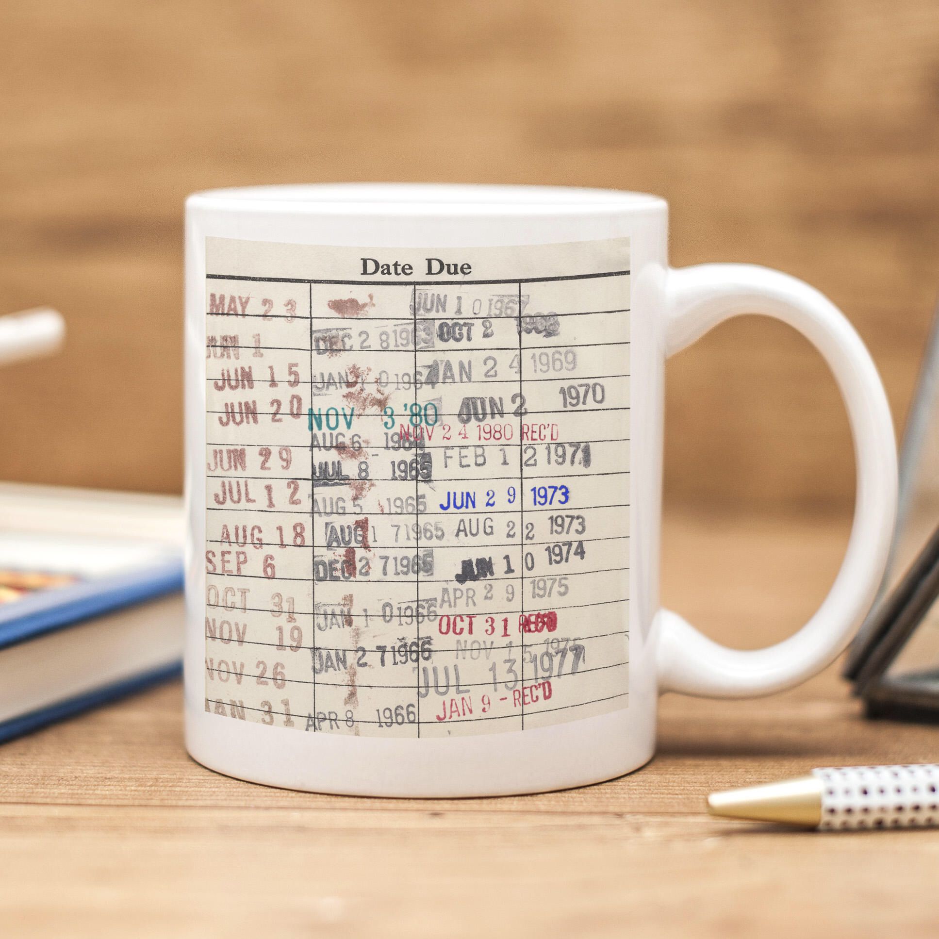 White-colored and dated library card mug on a wooden tabletop with a pen and a book around on a wooden tabletop