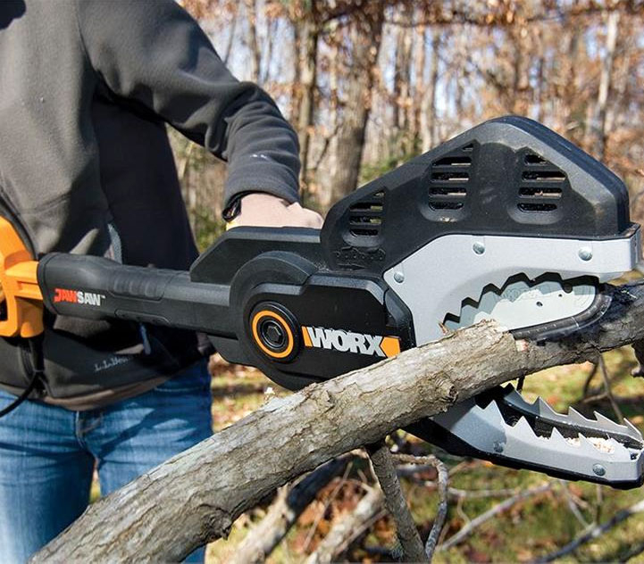 A man, wearing a black hoodie and jeans,  cutting a branch of tree with the help of black colored jaw saw