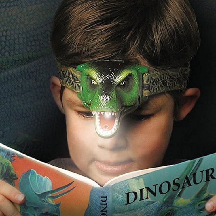 A boy wearing a green dinosaur head flashlight while reading a dinosaur book