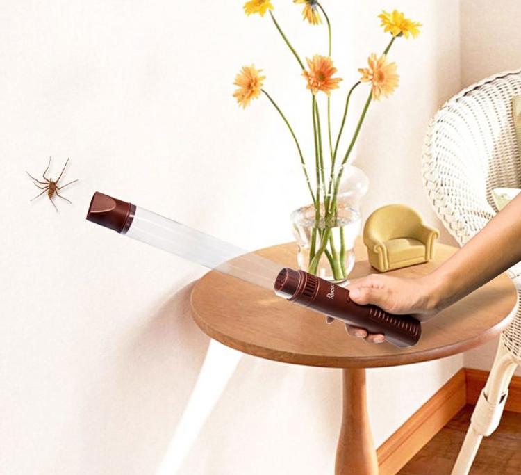 A hand is holding a brown and transparent-colored spider catching vacuum beside a wooden table with a glass vas, flowers, and a mini toy sofa 