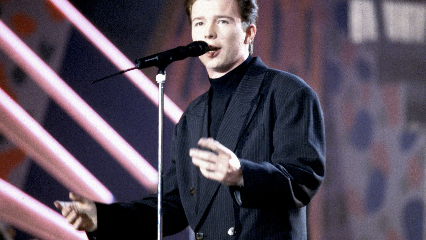 Rick Astley wearing a black outfit and singing on stage