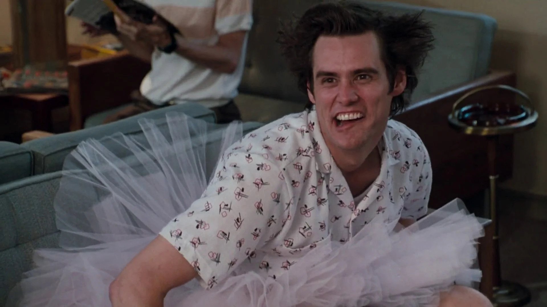 Ace Ventura wearing tutu costume and sitting on a sofa with a quirky face expression