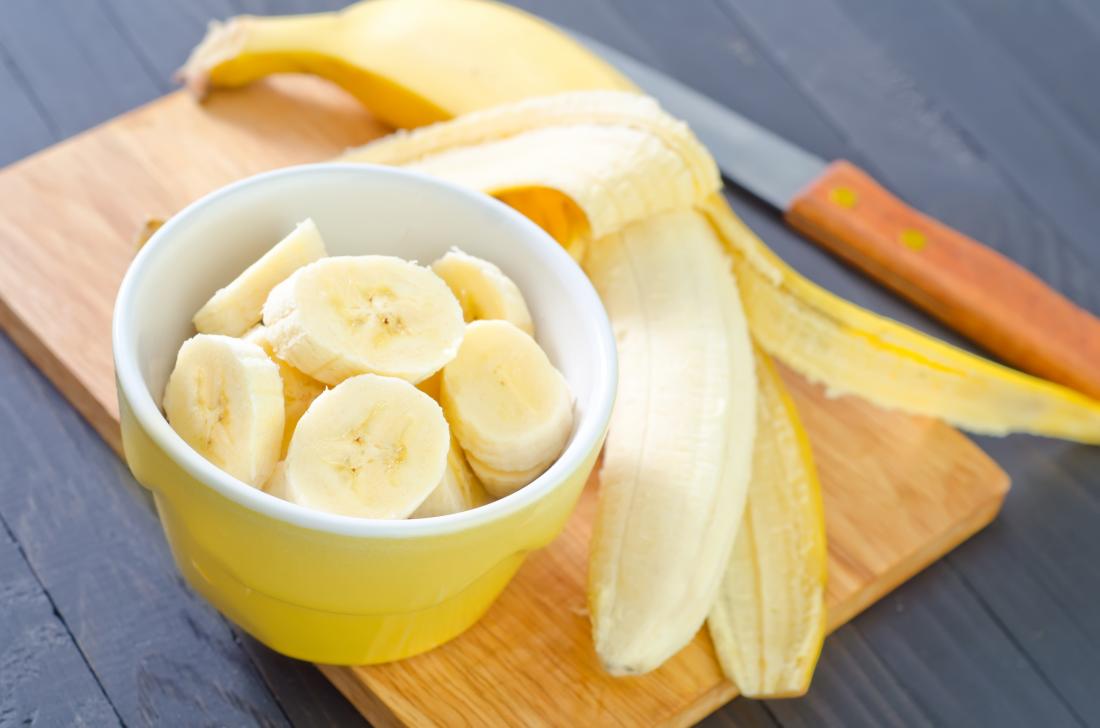 Banana chops in a yellow-colored bowl and on the chopping board is a peeled banana