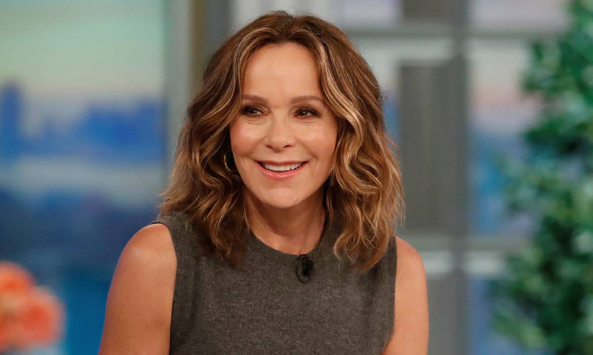 Jennifer Grey is wearing a gray sleeveless outfit and is smiling somewhere