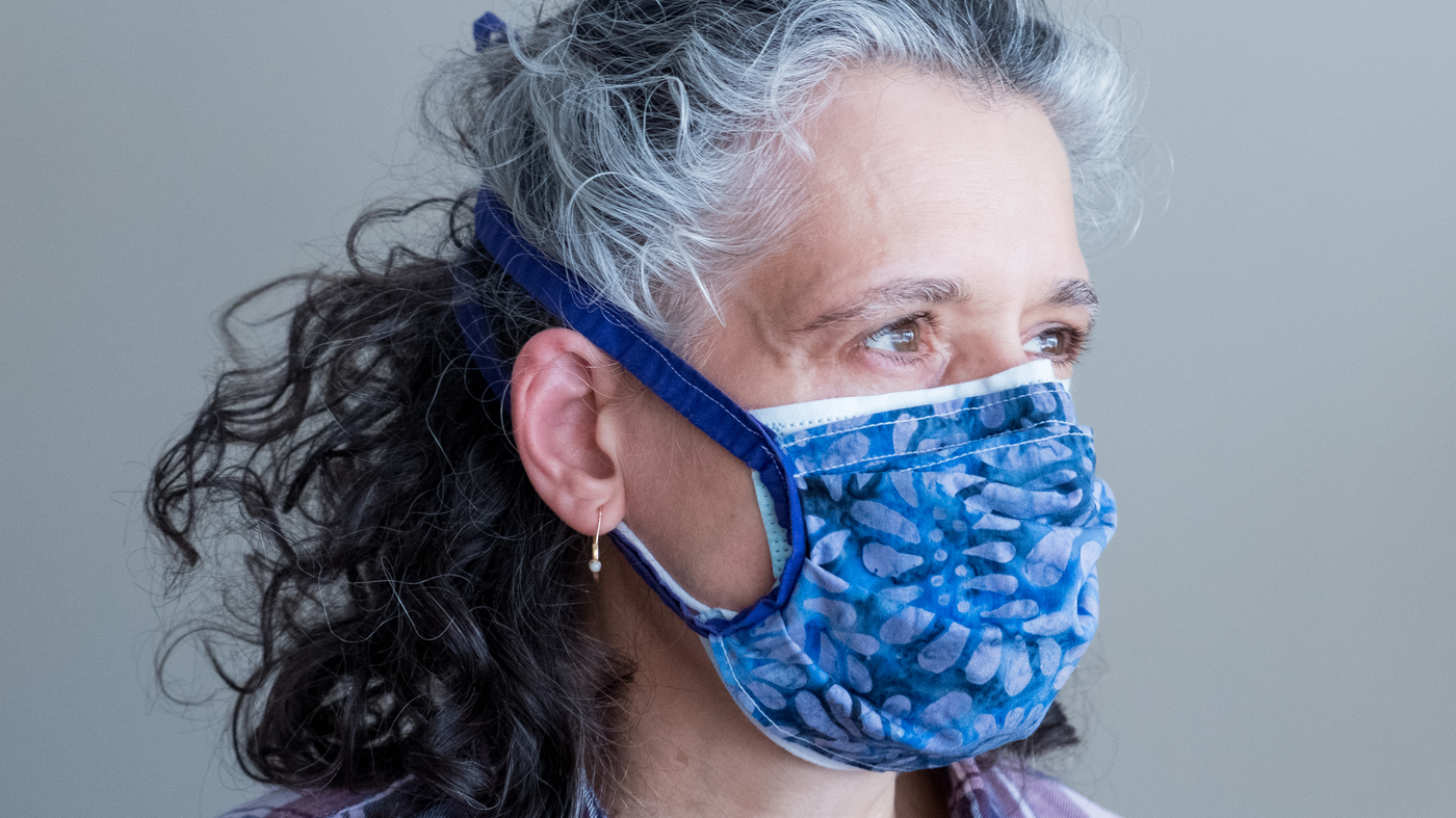 A woman having a gray hairs is wearing a medical facemask and layered with a cloth mask