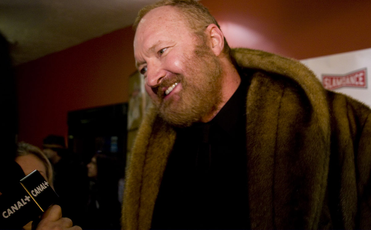 Randy Quaid is wearing a thick brown jacket and is listening and looking at the press