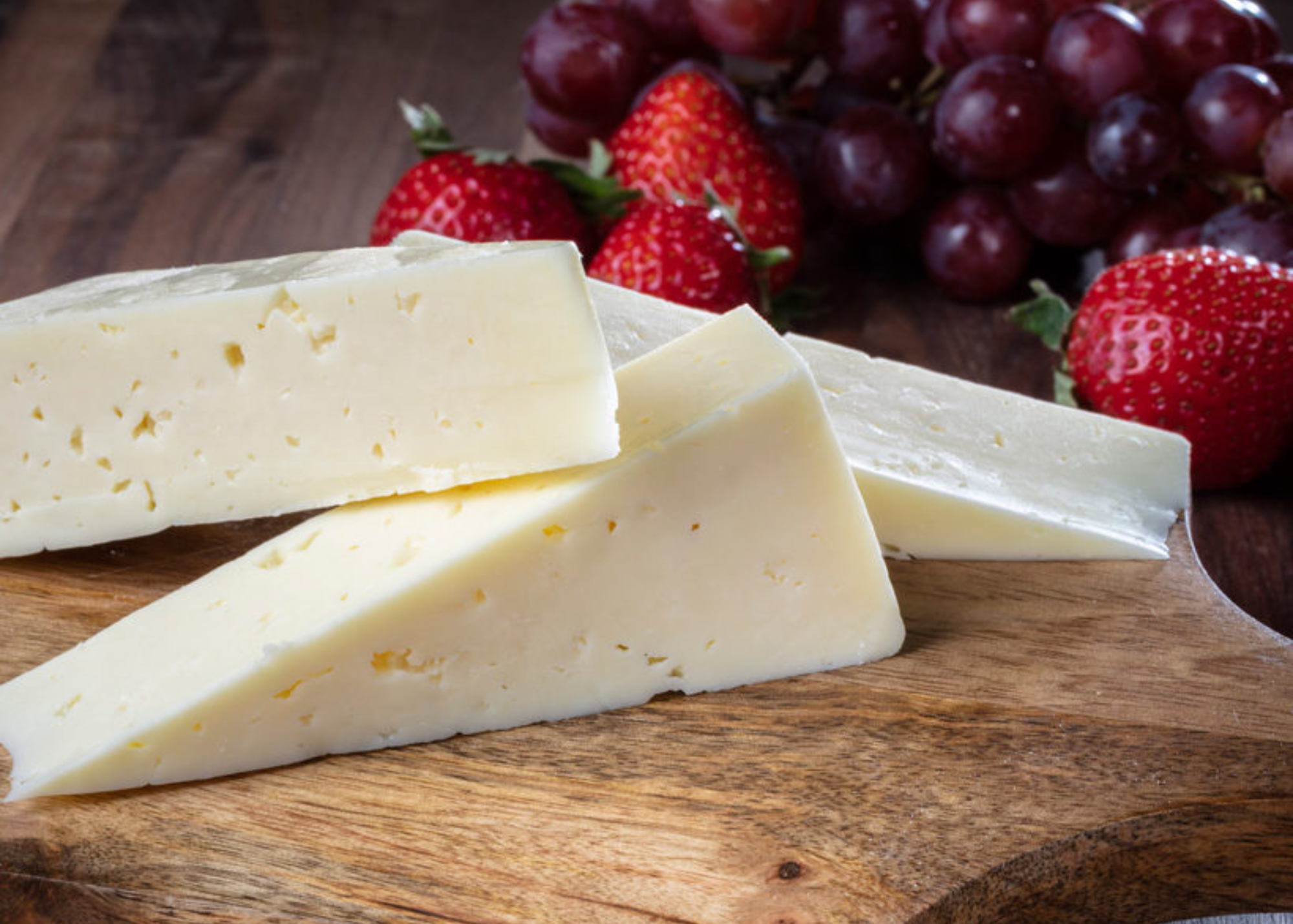 Three thick slices of havarti cheese are placed on a wooden board, with strawberries and grapes behind it