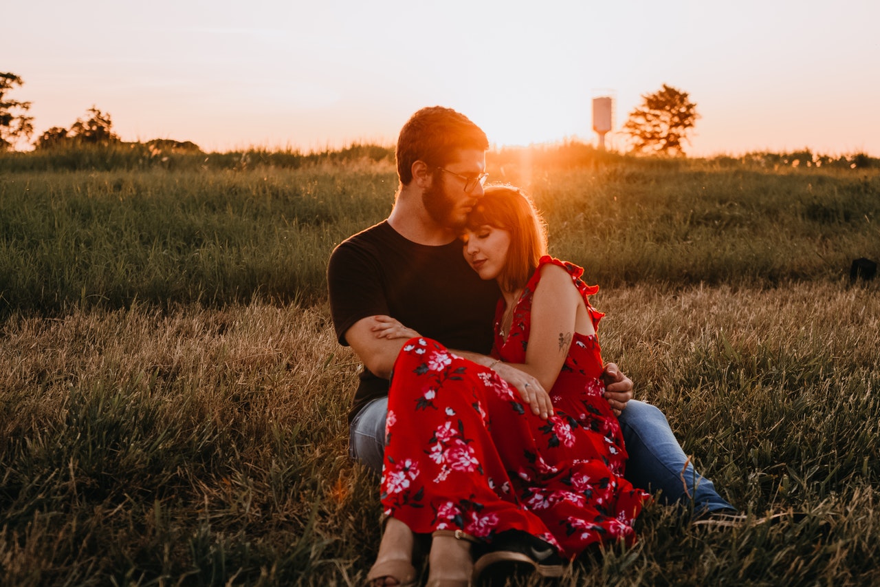 Man In Black Crew Neck T Shirt Sitting On Grass Field Beside Woman In Red Floral Dress