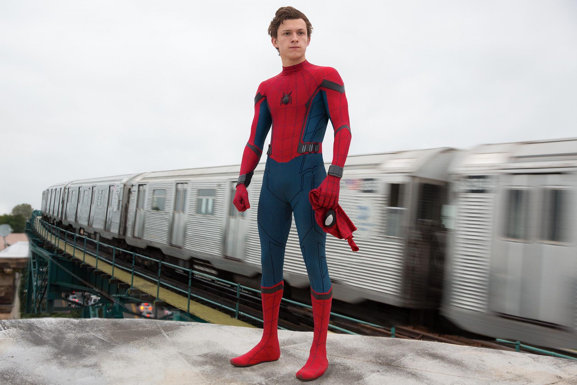 TomHolland is uncovered its entire headmask and is standing in front of a moving train on a railroad track