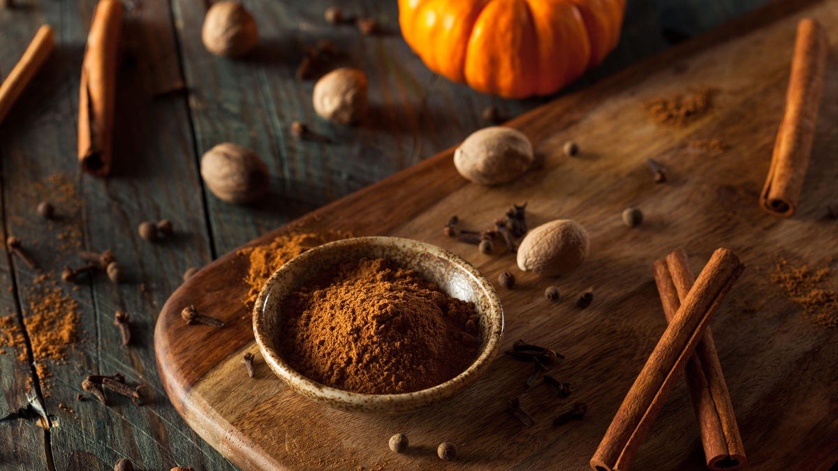 Pumpkin spice in a wooden bowl near cinnamon sticks, nutmeg, clove, and pumpkin on a wooden surface