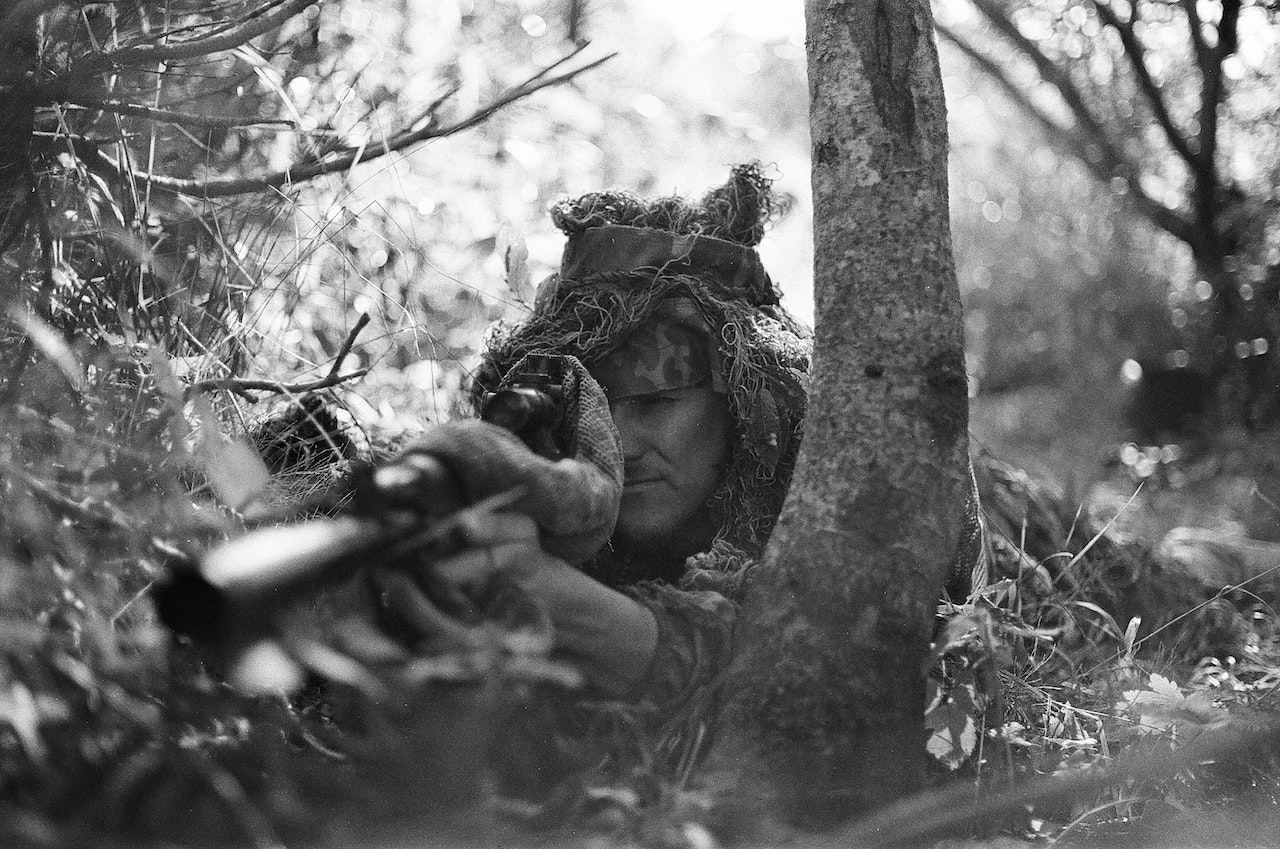 A sniper lying on his stomach in the forest and aiming his gun at something