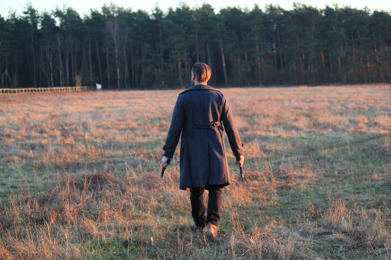 A Man In Coat Walking In The Field While Holding A Gun
