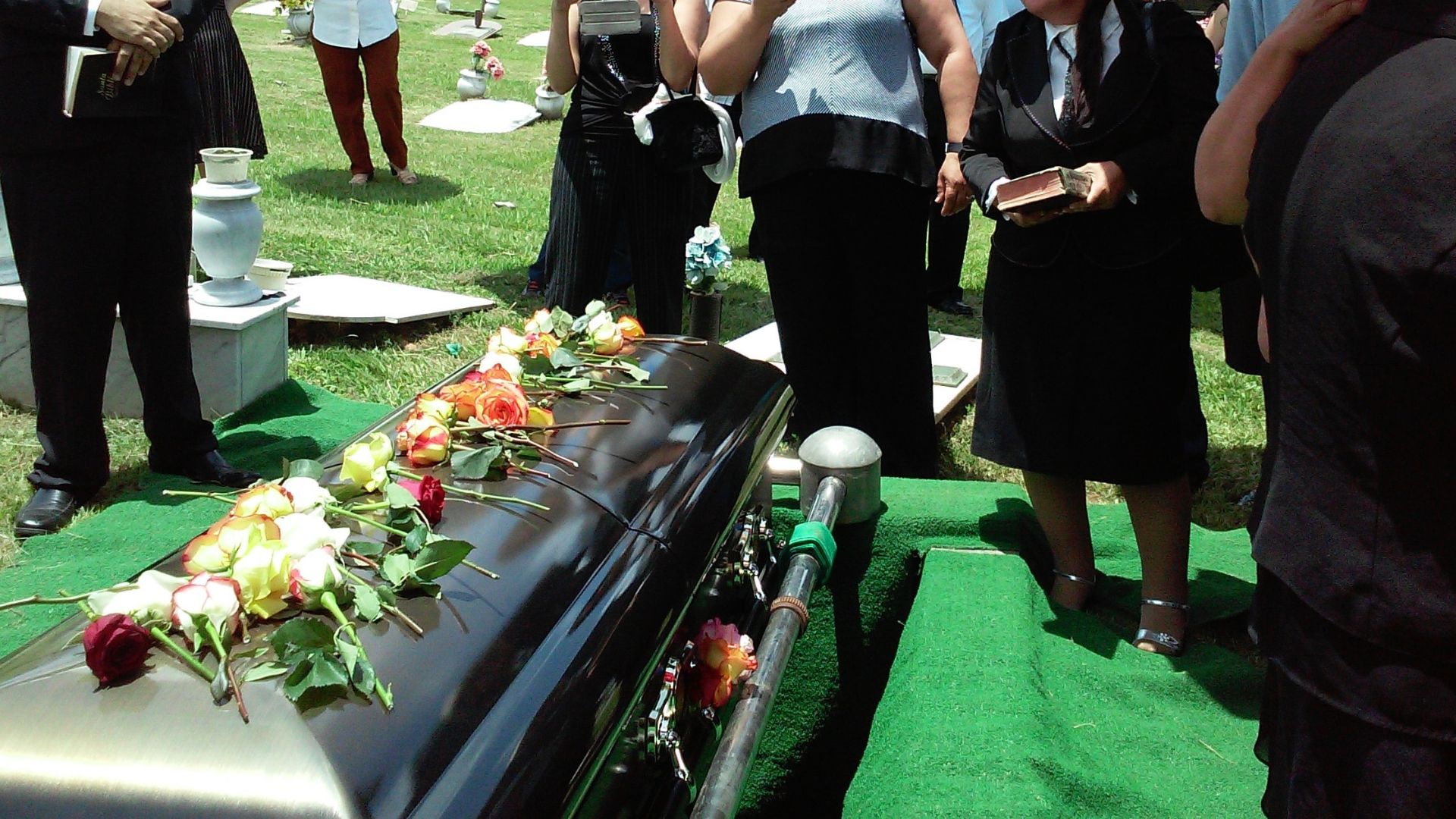 Roses on top of a black casket surrounded by grieving people