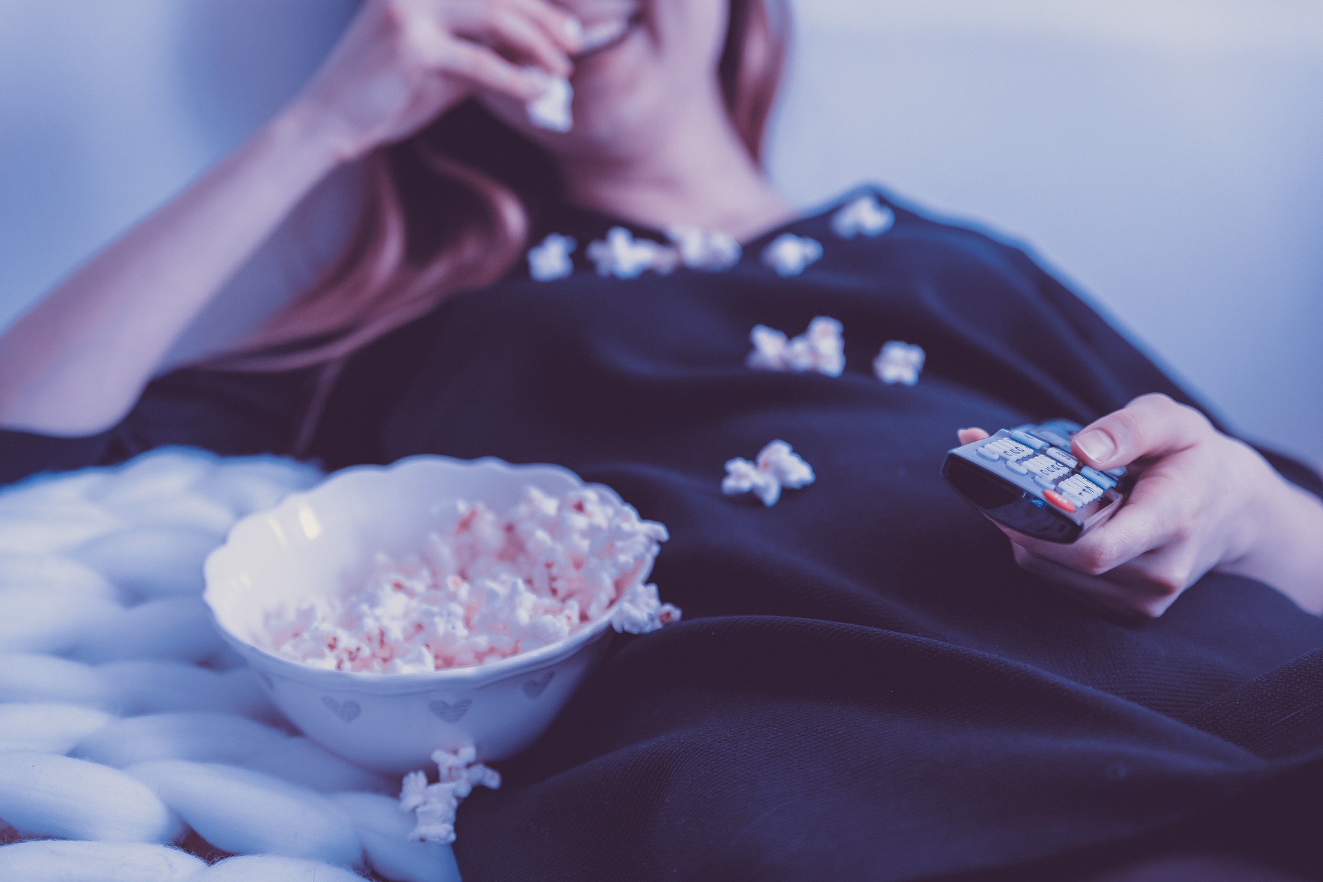 A woman is eating popcorn in bed while holding a TV remote