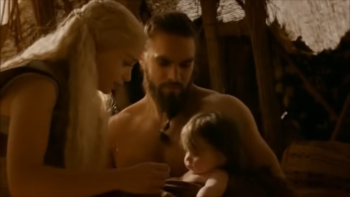 Daenerys reaching out to her unclothed son clutched by a sitting Drogo inside a tent